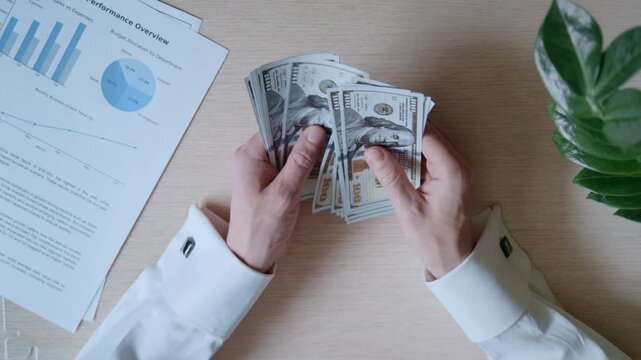Businessman counting a stack of hundred-dollar bills at his office desk with financial reports