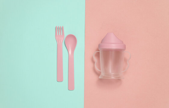 Drinking bowl and plastic fork and spoon for baby on a blue and pink background. Top view