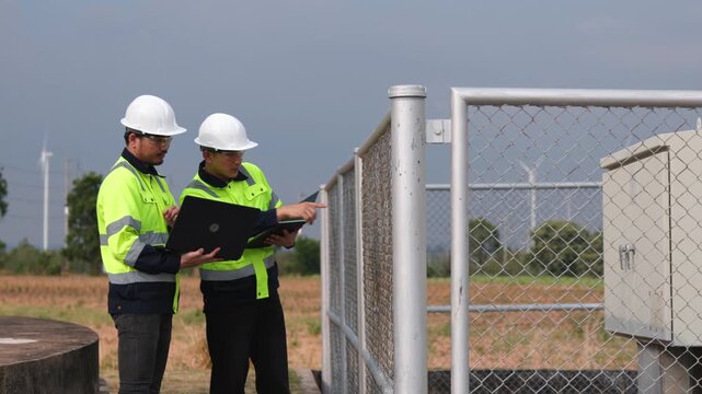 Professional Asian windmill engineers watching drone video feed on screen outdoor near grid. Two industrial safety technicians reviewing clean electricity generator strategy.