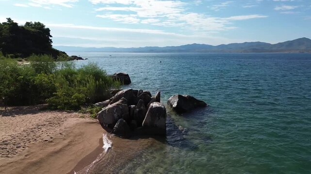 Drone orbits rock formation straddling water and sand on coast on a sunny day on Likoma Island in Lake Malawi