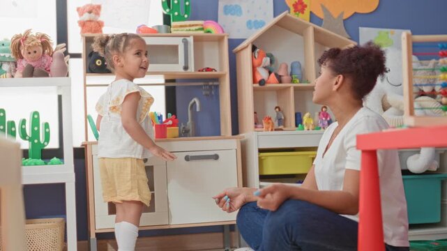Woman kneeling watching little girl open toy kitchen cabinet, fingers on handle near shelves and plush toys in a preschool building; nurturing care.