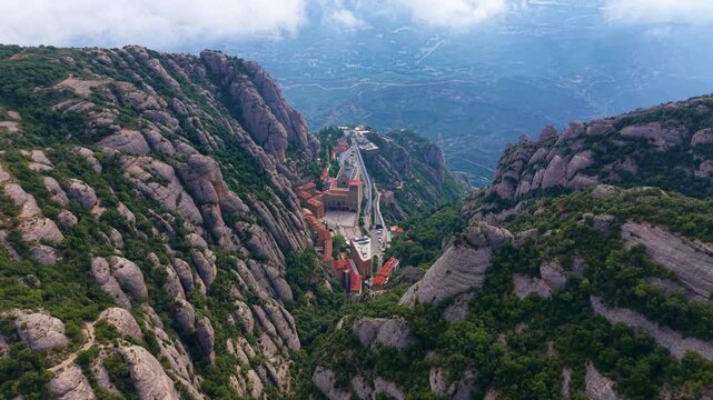 Aerial view down the valley of Montserrat monastery complex wedged between dramatic vertical conglomerate rock formations, with misty Catalan plain stretching into the distance, Spain