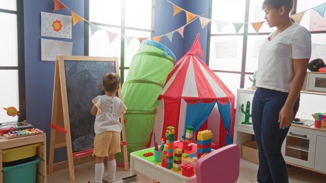 Girl erases large chalkboard with green sponge while standing at easel in building playroom surrounded by toys; concentration early learning.