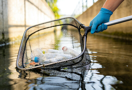 A person wearing blue gloves uses a net to remove discarded plastic bottles from a narrow waterway.
