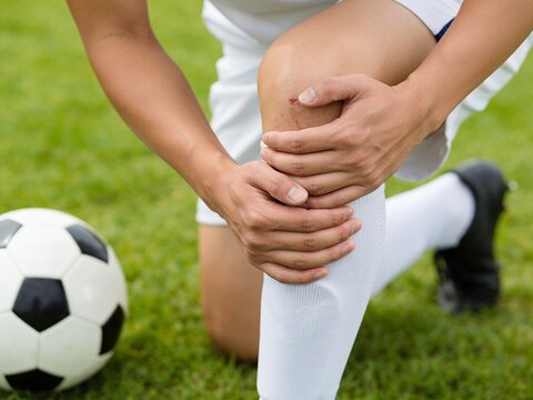 A soccer player kneels on the grass with hands clutching a painful, scratched knee during a match.