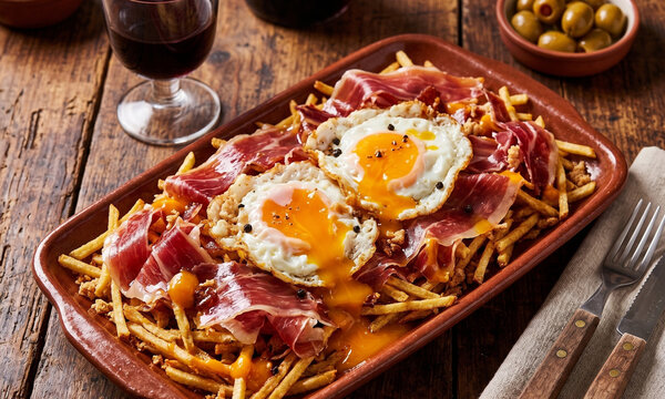 Huevos rotos tradicionales espa&ntilde;oles con jam&oacute;n ib&eacute;rico y patatas fritas paja en plato de barro