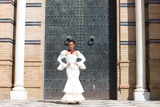 A beautiful woman dancing flamenco in front of a giant door in Seville, Spain. She is wearing a traditional white dress with ruffles and has red flowers.Flamenco, a UNESCO Intangible Cultural Heritage