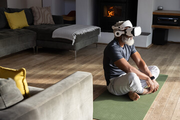 Obraz premium Senior African American man sitting on green yoga mat wearing VR headset near fireplace, copy space