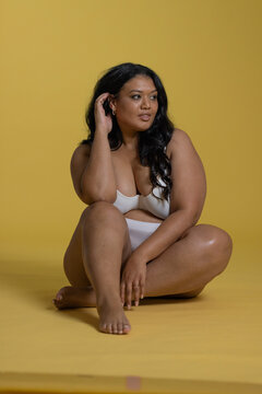 Mid-adult woman sitting cross-legged in studio on yellow backdrop in white underwear touching hair