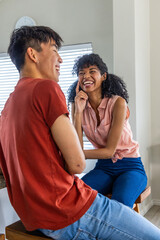 Naklejka premium Diverse couple sitting on wooden stools wearing red tee and pink blouse laughing by window blinds