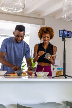 African American couple making breakfast bowls at white island, filming on phone tripod