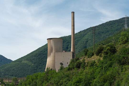 Abandoned thermal power plant with cooling tower and smokestack surrounded by forested mountains. Industrial infrastructure left unused in a natural landscape, illustrating industrial decline and ener