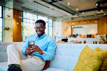 Young businessman relaxing on a sofa, smiling while interacting with his mobile phone in a modern...