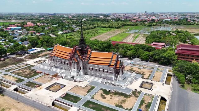 Impressive drone view of a traditional Buddhist temple under construction in Ang Thong, Thailand. Features a multi-tiered orange roof and a tall central spire wrapped in red scaffolding.