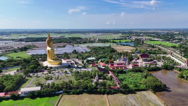 Spectacular wide drone shot of the Great Buddha of Thailand in Ang Thong. Features the country's biggest golden statue overlooking expansive temple grounds, green rice fields, and rural landscapes.