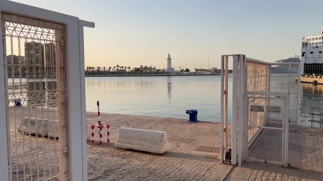 View of the calm harbor water and the historic La Farola lighthouse through an open security gate at Muelle Uno in Malaga, Andalusia, Spain.