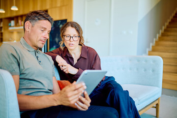Naklejka na ściany i meble Man and woman relaxing at home, looking at a digital tablet screen together and having a casual conversation about content