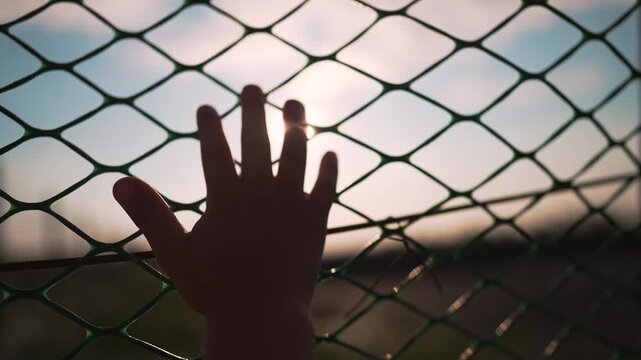 Hand reaching through chainlink fence at sunset with fingers spread wide. Person touches fence barrier with hand. Silhouette shows hand on chainlink mesh. Fingers reach through wire fence.