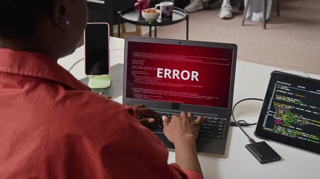Over the shoulder view of African American female developer typing on laptop and encountering red system error screen. Footage represents cybersecurity threats, software bugs, or IT support services