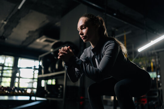 Athletic woman performing deep squat exercise with focused intensity, training legs and glutes in modern gym bathed in dramatic, motivating lighting during powerful workout session.