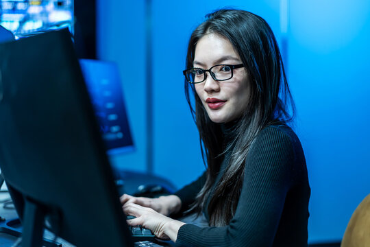 Focused Asian female software engineer in a modern dark office, working on computer code. Professional programmer developing technology solutions in a high-tech illuminated workspace.