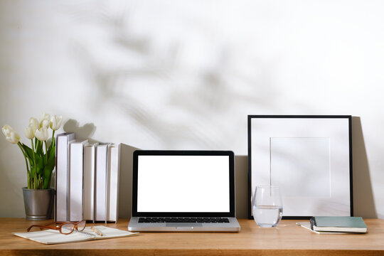 Minimal home office desk with blank laptop screen, white tulips, books, notebook, eyeglasses, and framed mockup against a bright wall.