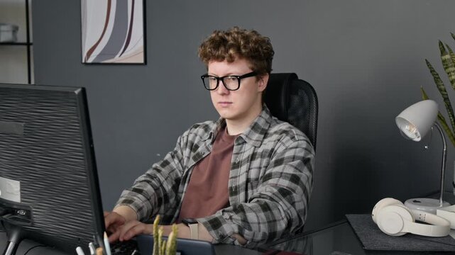 Caucasian young male programmer wearing glasses typing on computer keyboard at office desk. Clip for technology, software development, and remote work commercial videos
