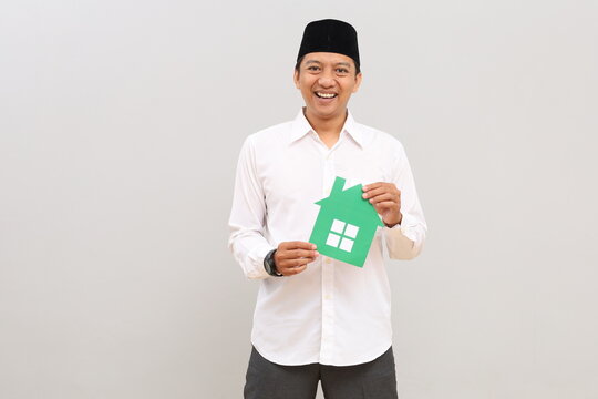 Asian Muslim man holding a small paper house image, posing in front of a gray background wall with empty space for promotional content or design.