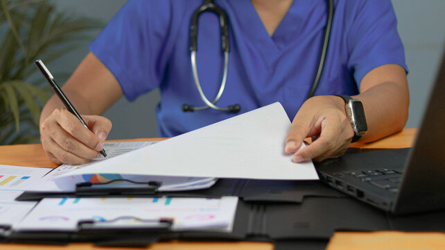 Healthcare professional reviewing medical documents and signing reports at a modern clinic desk. Doctor managing patient records, healthcare administration, medical paperwork, and digital systems.