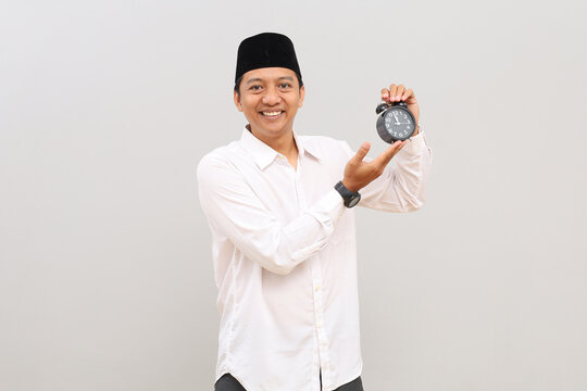 Asian Muslim man showing alarm clock, Ramadan fasting month concept, smiling looking at camera, standing isolated on grey background.