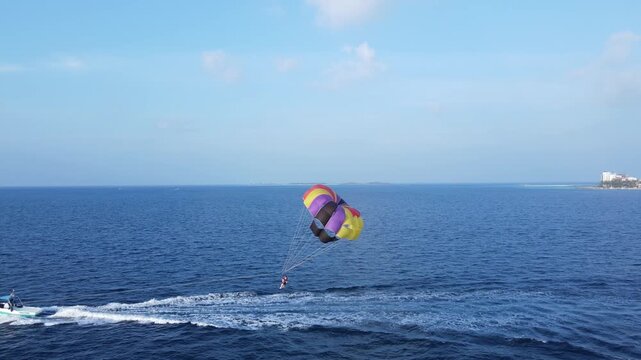 Drone view of couple people parasailing with a colorful parachute pulled by a speedboat over blue tropical ocean. The scene highlights adventure, summer vacation, and water sports.