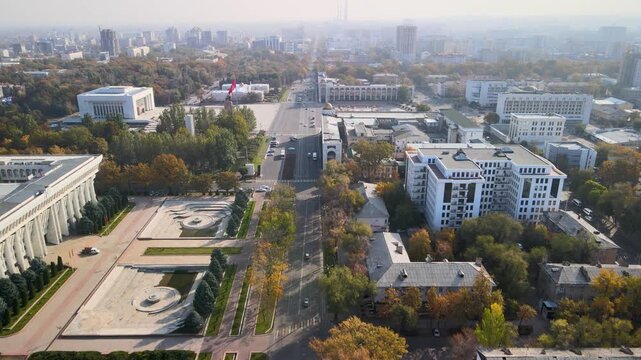 Drone flyover along a central avenue in Bishkek, Kyrgyzstan, passing a city square with a national flag and surrounding urban blocks.