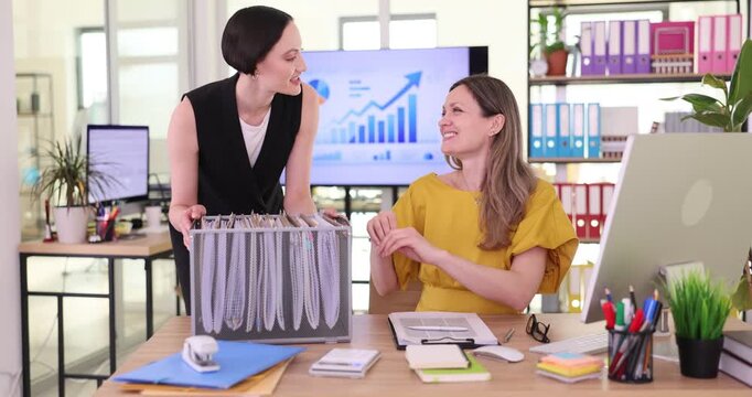 Businesswoman in formal attire brings work folders in box to secretary sitting at desk. Woman office employee studies important documents at table