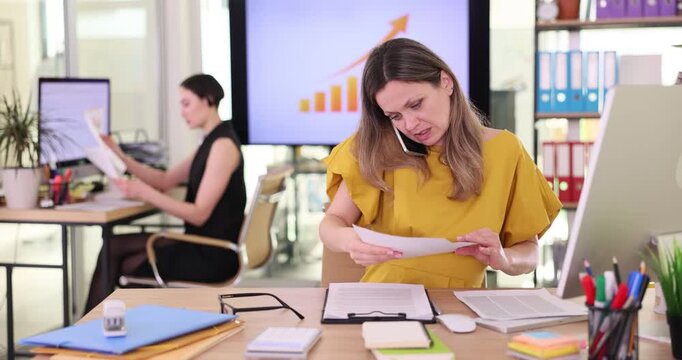 Concentrated woman manager talks on phone searching for important document at table. Office worker discusses important business matters by telephone