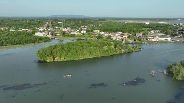 Drone video capturing a small motorboat sailing on the river estuary next to a green island and pedestrian bridge in the coastal resort of Anaklia, Georgia, with the sun reflecting on the water.