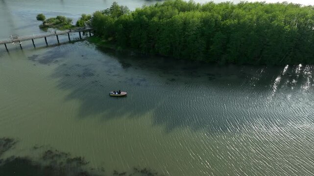 Drone video capturing a small motorboat sailing on the river estuary next to a green island and pedestrian bridge in the coastal resort of Anaklia, Georgia, with the sun reflecting on the water.