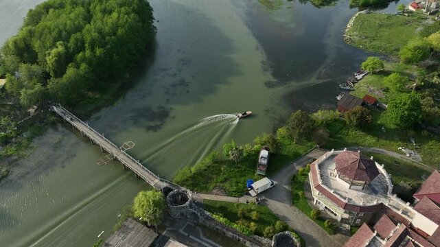 Drone video capturing a small motorboat sailing on the river estuary next to a green island and pedestrian bridge in the coastal resort of Anaklia, Georgia, with the sun reflecting on the water.