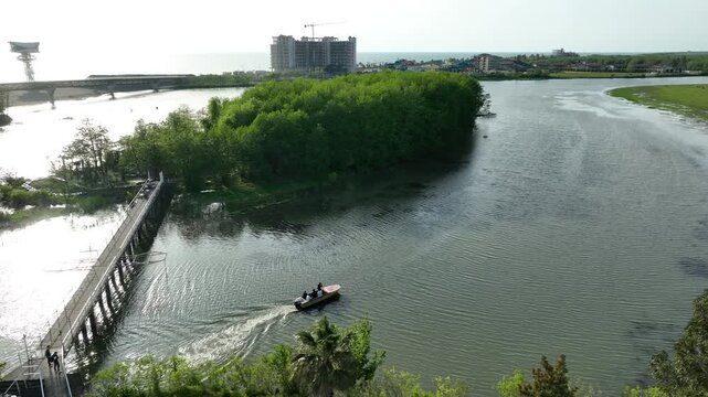 Drone video capturing a small motorboat sailing on the river estuary next to a green island and pedestrian bridge in the coastal resort of Anaklia, Georgia, with the sun reflecting on the water.