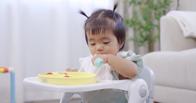 Asian toddler girl sitting on feeding chair holds banana slice bites gently, learning self feeding skill, soft daylight brightens home interior, playful breakfast scene, early development curiosity