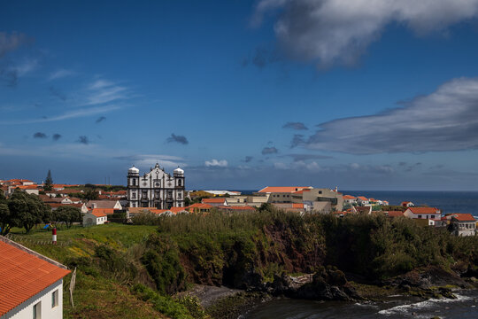 Vista panor&aacute;mica de un pintoresco pueblo, Santa Cruz das Flores, con casas de tejados naranjas y una gran iglesia hist&oacute;rica sobre un acantilado verde junto al oc&eacute;ano Atl&aacute;ntico bajo un cielo nublado.