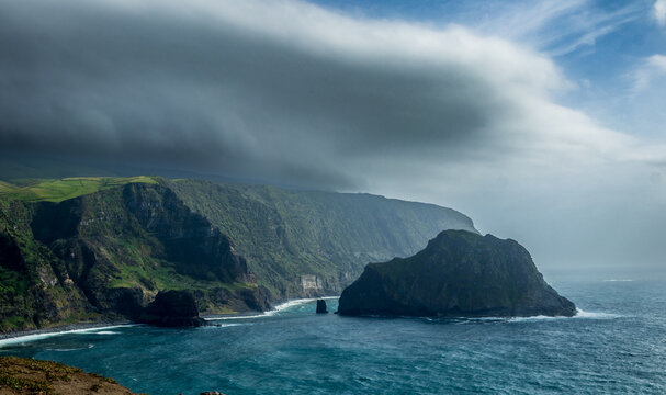 Un paisaje costero en la isla de Flores, Azores, con acantilados verdes cubiertos de niebla y un gran islote de piedra en el mar, bajo un cielo amenazante con densas nubes oscuras.
