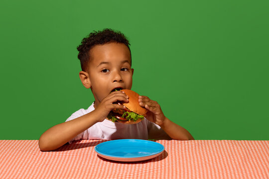 Young boy biting hamburger on green background with colorful plate. Concept of burger bite, kids menu advertising, childhood treat, and colorful food poster.