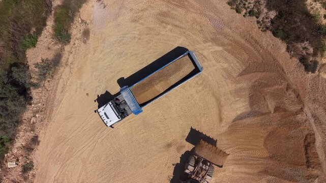 Top-down view of a bulldozer pushing sand into a semi-truck trailer at a sandy construction or quarry site surrounded by sparse vegetation