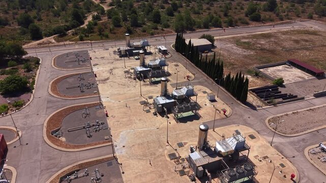 Overhead view of a gas compressor station with industrial equipment, pipelines, valves, and mechanical units surrounded by dry terrain and cypress trees