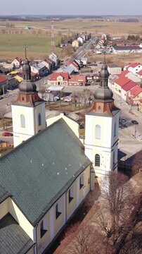 Vertical screen Aerial View Stawiski Church Roof And Twin Towers, Small Town Backdrop With Red Rooftops, Distant Fields And Clear Sky, Warm Daylight Highlighting Roof Texture And Tower Domes
