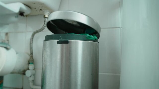 Dirty public restroom with waste bin near toilet stall. Discarded paper entering trash container inside neglected bathroom facility. Sanitation problems, restroom hygiene, public cleaning services