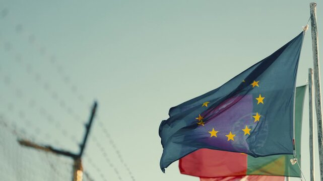European flag waves in the wind near a border with a glimpse of another country s flag and a fence in the background