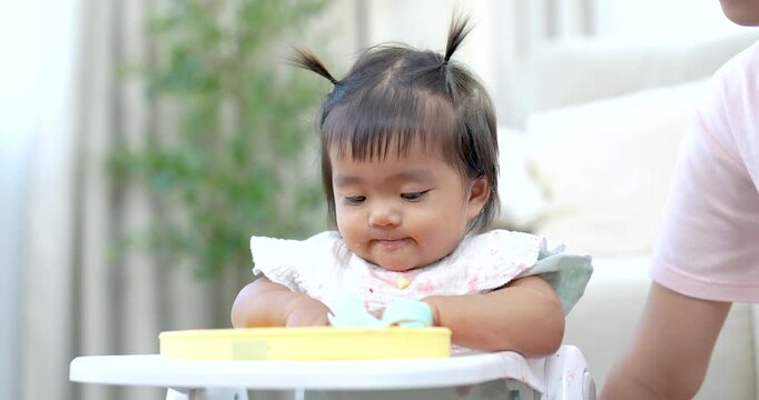 Asian toddler girl sitting on feeding chair playing with spoon food tray, touching vegetables curiously, smiling with joy, learning self feeding skills, indoor family lifestyle, natural daylight