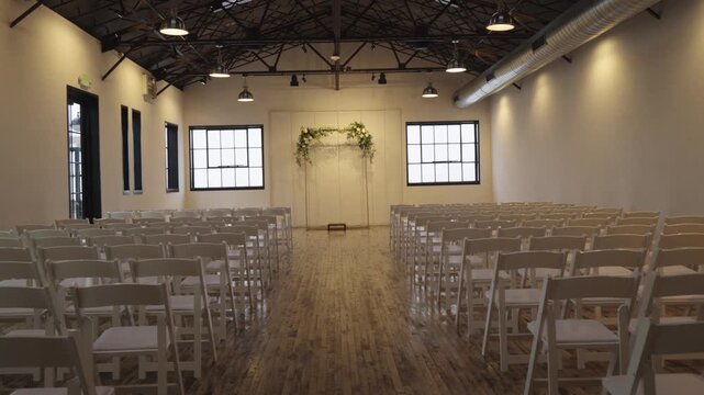 A modern event space with wooden floors features rows of white chairs facing a floral arch, ready for a gathering.