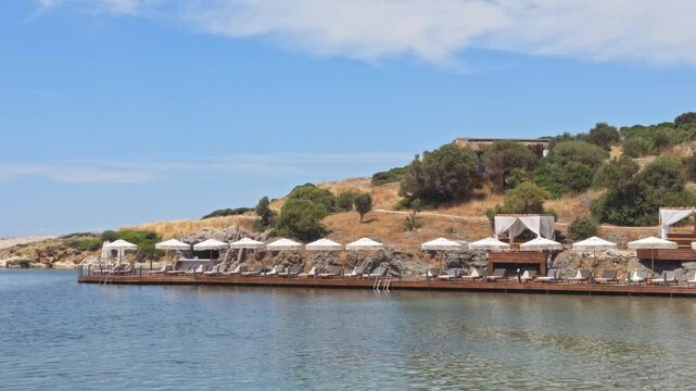High-quality 4K wide shot of an elegant wooden sun deck over the calm sea at a luxury beach resort. Features white umbrellas, loungers, and a seaside bar.